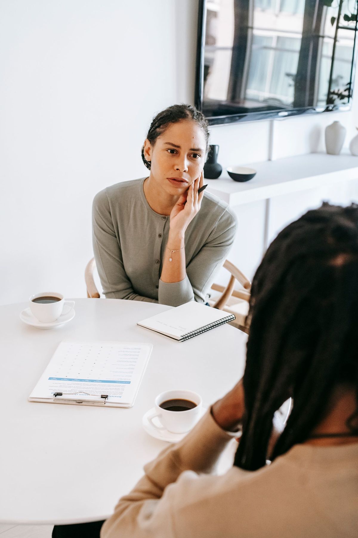 A counselor sits at a round white table with coffee cups, a notebook, and a clipboard, resting her chin on her hand as she listens closely to the person seated across from her.
