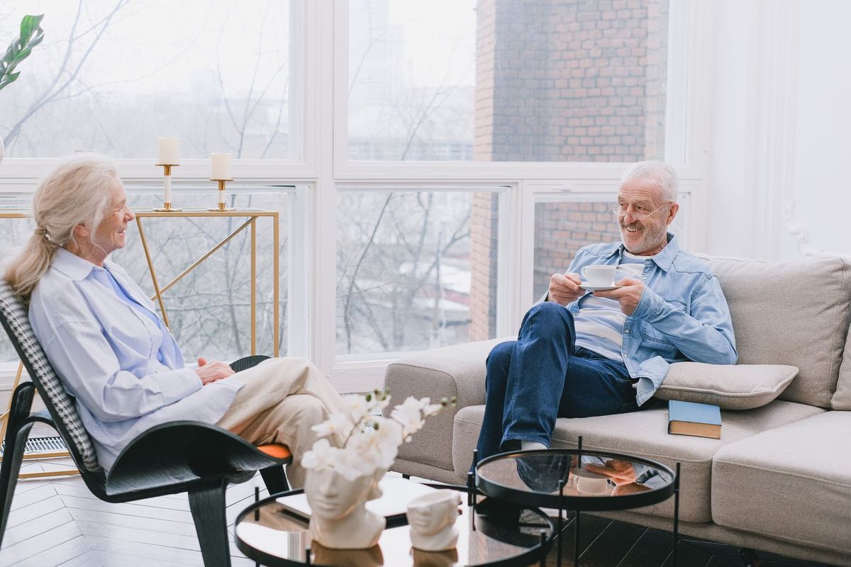 An older couple smiles and talks together in a bright living room with tall windows. The woman sits in an armchair and the man sits on a sofa holding a cup of coffee.